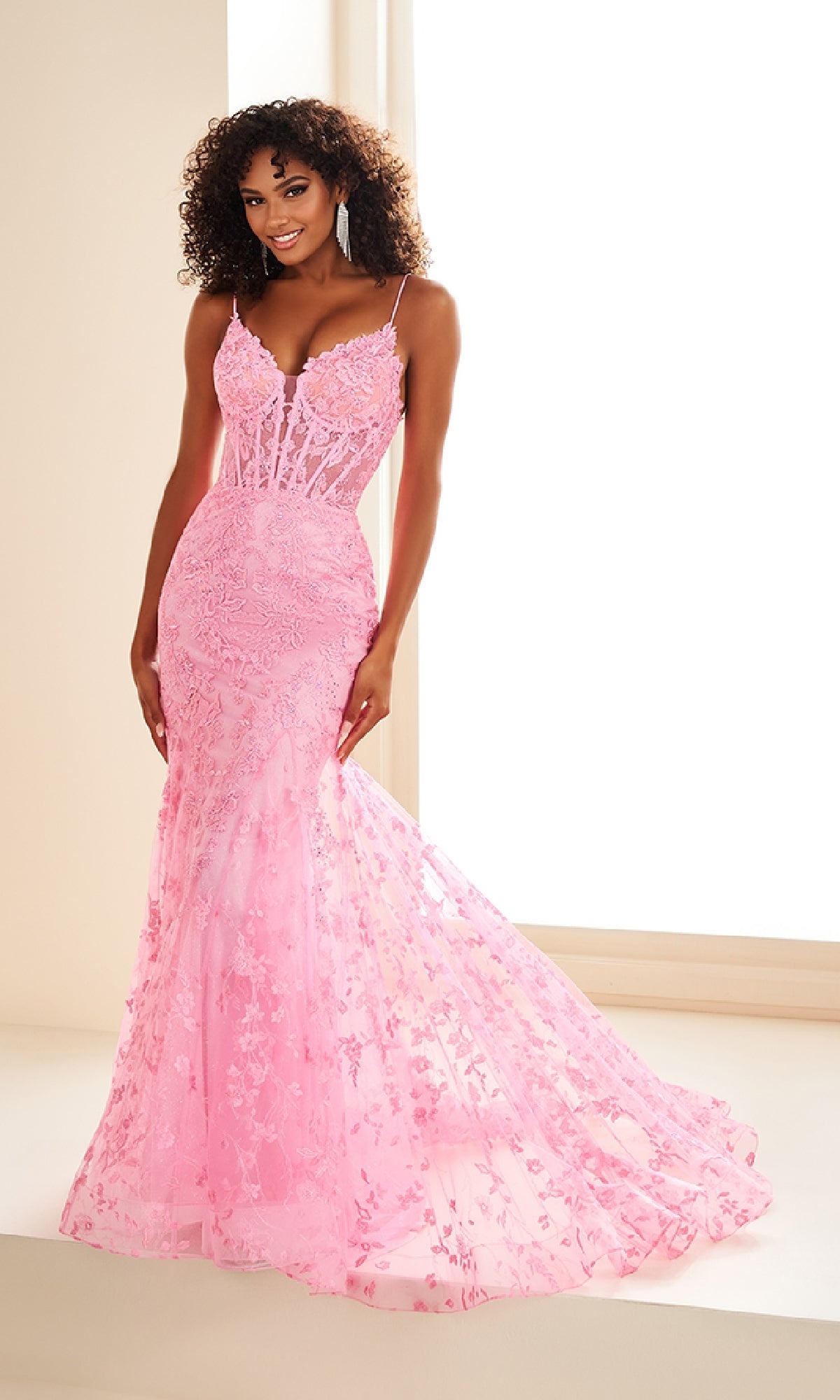 A woman stands indoors wearing the Ellie Wilde EW36008, a fitted, floor-length light pink prom dress with a sheer bodice, lace-up back, and floral embroidery.